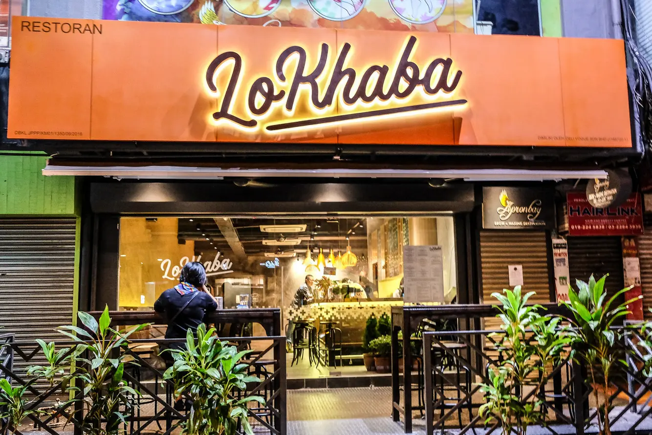 LoKhaba restaurant shopfront at night with backlit orange sign and railed pavement seating
