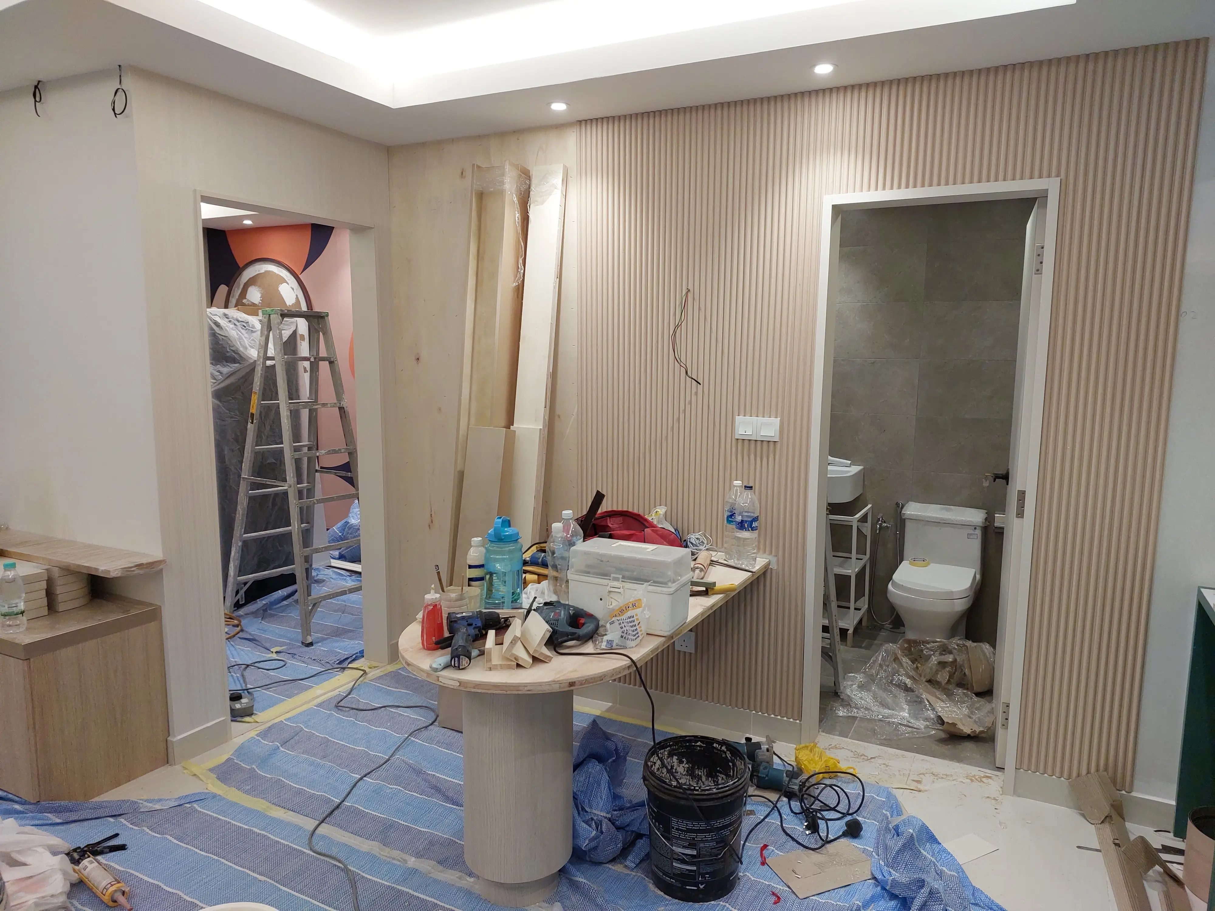 Fluted timber panelled room mid-fit-out, tiled washroom beyond, stepladder, tools and blue floor sheeting