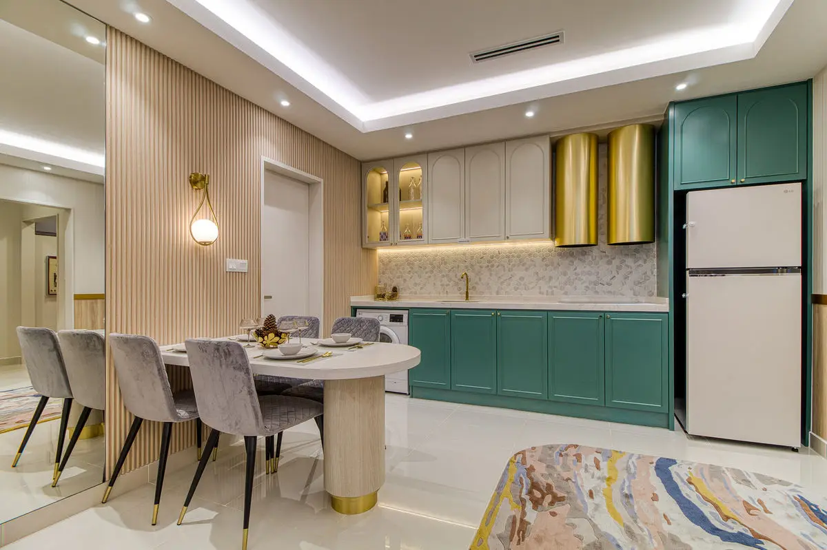 Dining table with velvet chairs, and a green kitchen with brass-trimmed cabinets behind