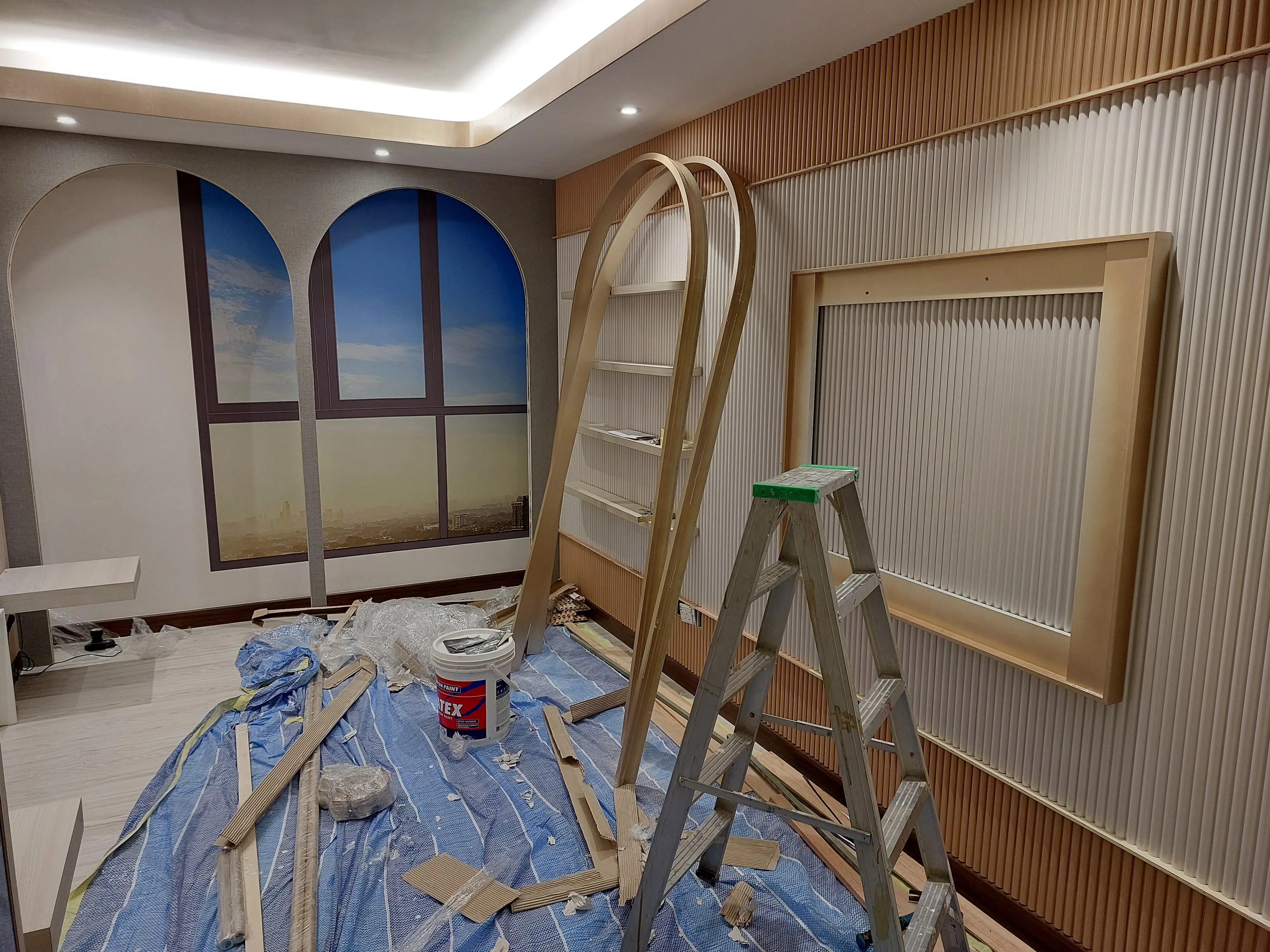 Room with painted arched window murals, fluted walls, open shelving, ladder and paint pail on sheeting