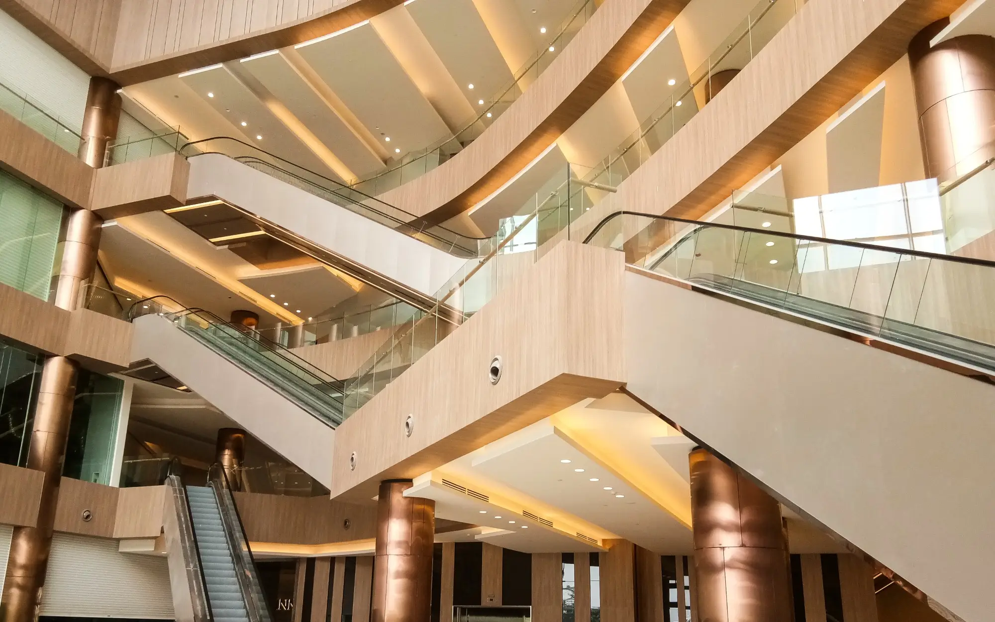 Mall atrium of crossing escalators, timber-clad soffits, bronze columns and warm cove lighting