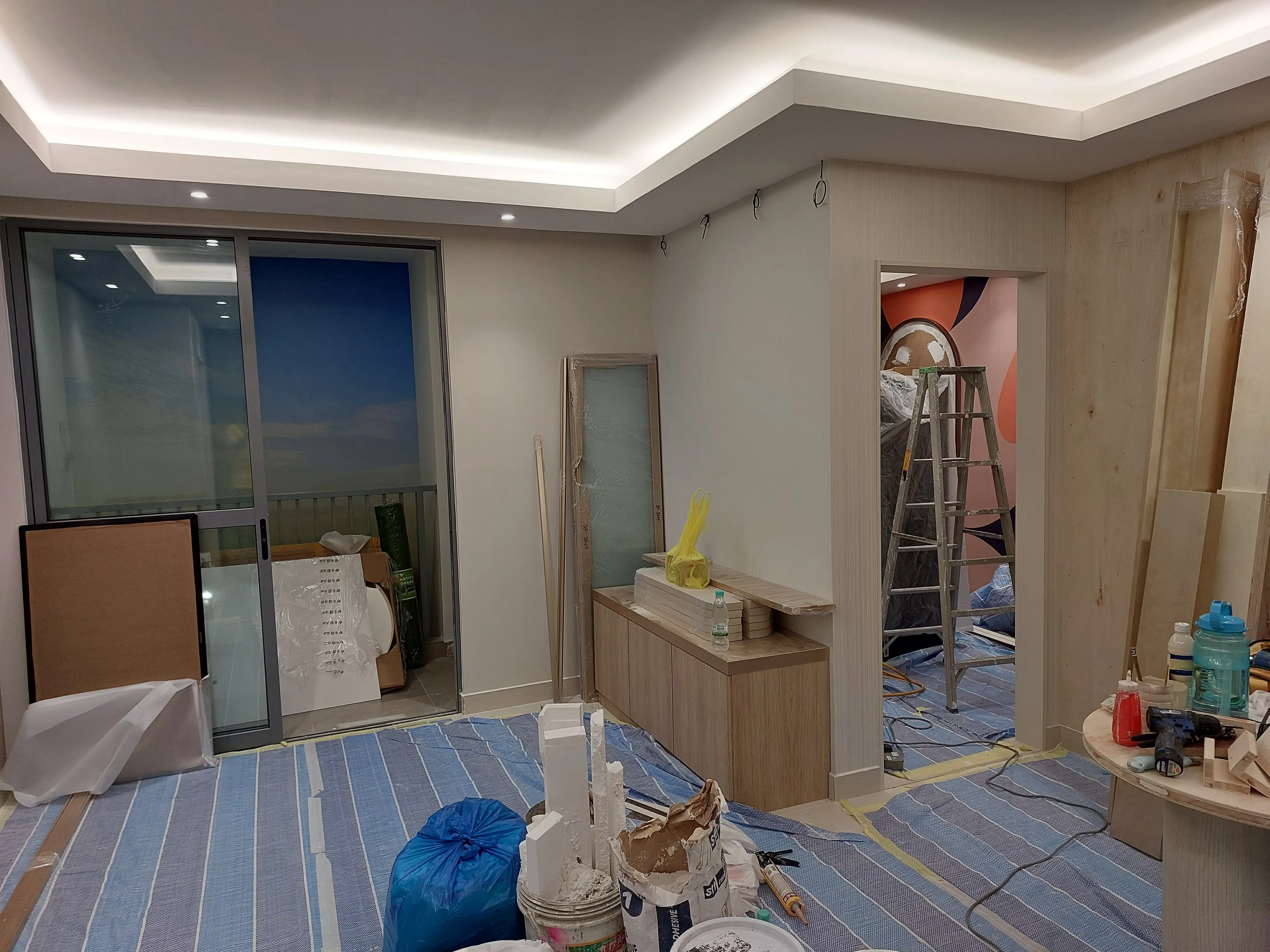 Living space mid-fit-out: glazed balcony door, oak sideboard, plaster bags, ladder and coral mural beyond