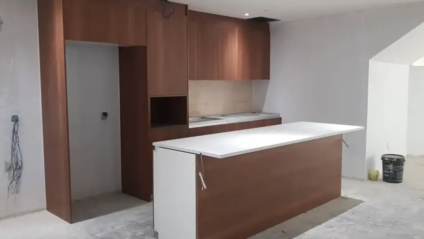 Bare-plaster kitchen shell with dark veneer tall units, white island worktop and exposed wiring